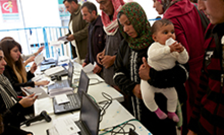 Photo of people sitting in front of laptops and lines of users in front of them demonstrating a sort of registration process