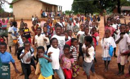 A joyful group of Zambian children, dressed in colorful clothing, gather outdoors near a school building in a rural area. They smile and wave energetically at the camera, with trees and simple structures in the background.