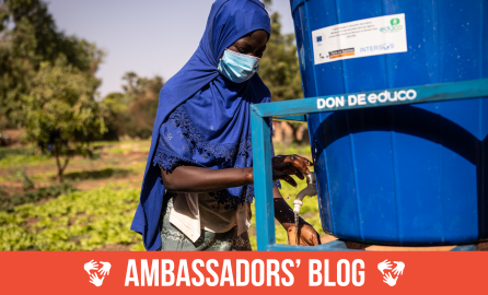A girl in a blue headscarf and mask fills a bucket with water at an outdoor water station, with greenery in the background