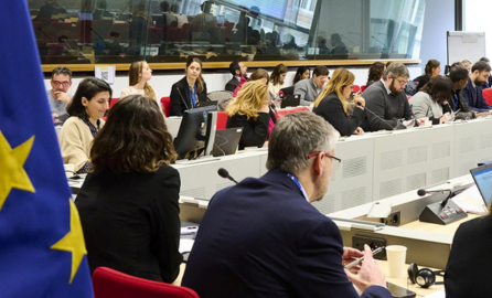 Meeting room with participants seated around conference tables, working on laptops and listening to the discussion. An EU flag is visible in the foreground, with more attendees and meeting equipment in the background.