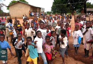 A joyful group of Zambian children, dressed in colorful clothing, gather outdoors near a school building in a rural area. They smile and wave energetically at the camera, with trees and simple structures in the background.