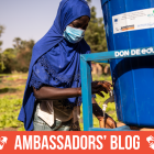 A girl in a blue headscarf and mask fills a bucket with water at an outdoor water station, with greenery in the background
