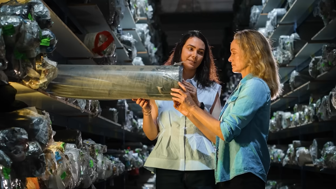 Amber Valletta, UNEP Goodwill Ambassador and Supermodel, seen with a worker in a denim factory, pulling a plastic tube in a deposit space where denim is packaged and stocked around them.